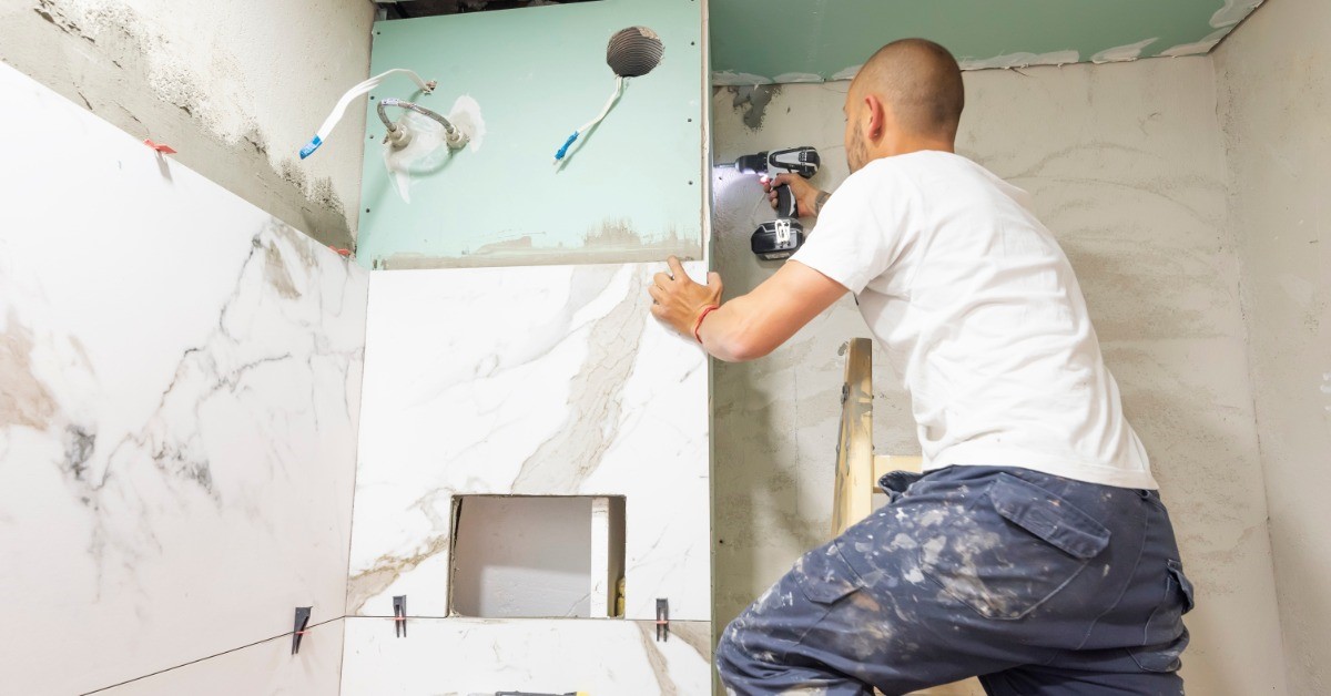 A man with a shaved head is standing on a step ladder and is using a drill on the side of a wall in a bathroom.