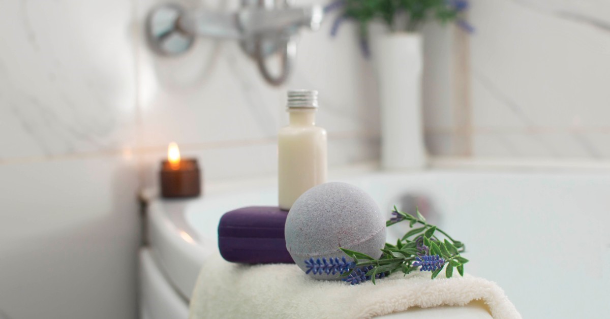 A tub has several items on its rim, including a candle, towel, bath bomb, and a container of a cream substance.