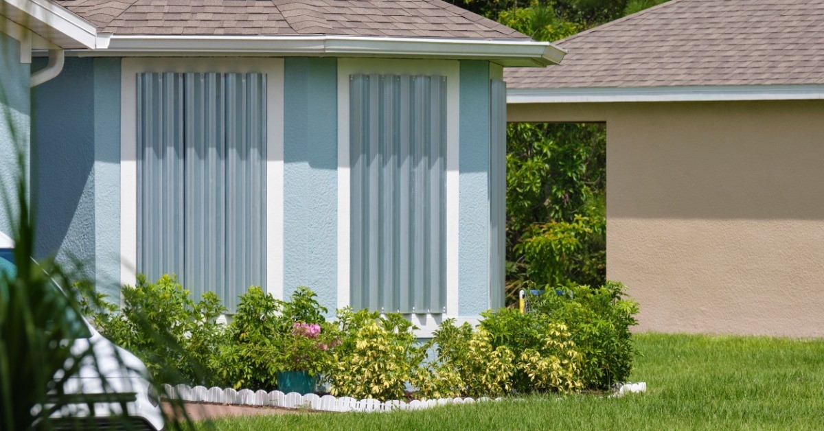 A residential home has its windows shuttered with metal. There is green foliage in the background.