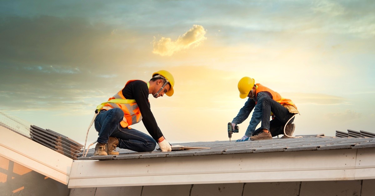 Two men are on a roof, both are wearing hard hats and high visibility vests. One man is holding a drill.