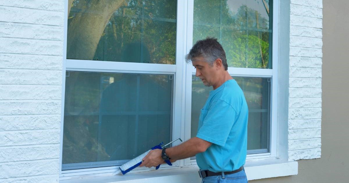 A homeowner applying caulk to the edges of his new hurricane windows with a caulk gun, ensuring a quality seal.