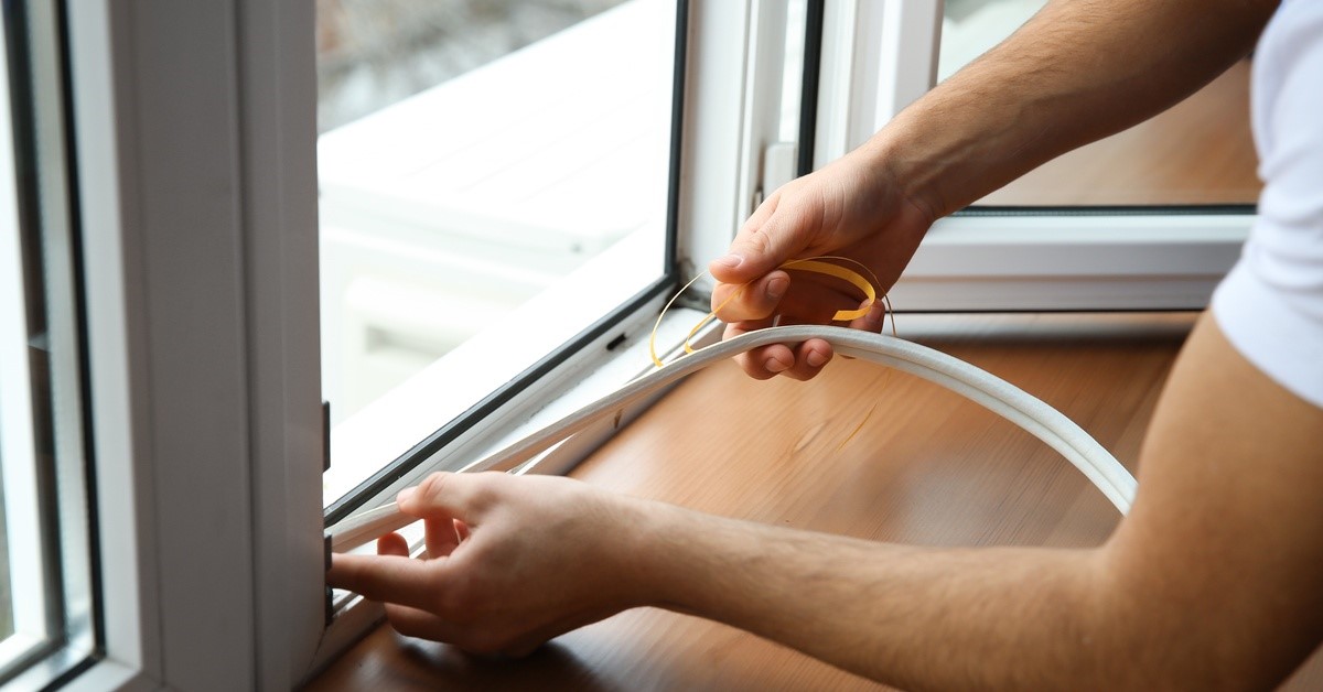 A person's hands holding weatherstripping seal materials as they work on a window. The window has a white frame.