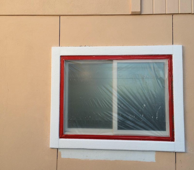A window with white and red trim covered with a plastic covering against a tan concrete wall. 