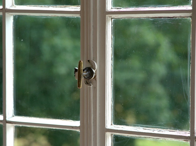 A close-up of a window with white trim on the grids with a gold lock piece. The glass is slightly worn and old looking.