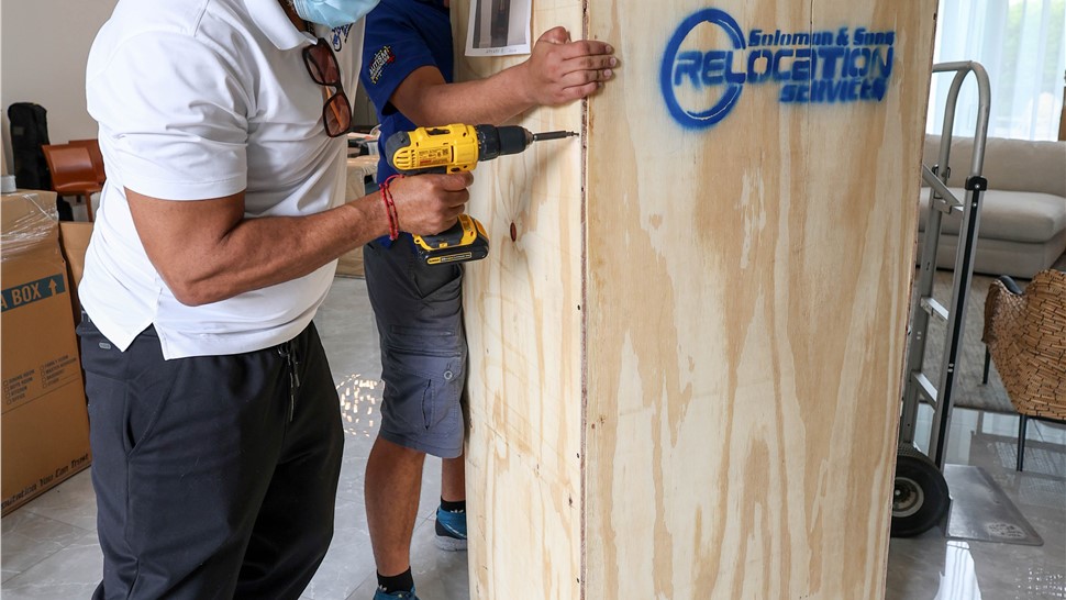 Movers using a drill to assemble a custom wooden crate inside a home.