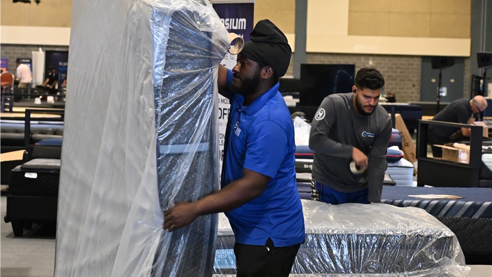 Mover in a blue uniform wrapping a large mattress or furniture piece in protective plastic inside a warehouse.