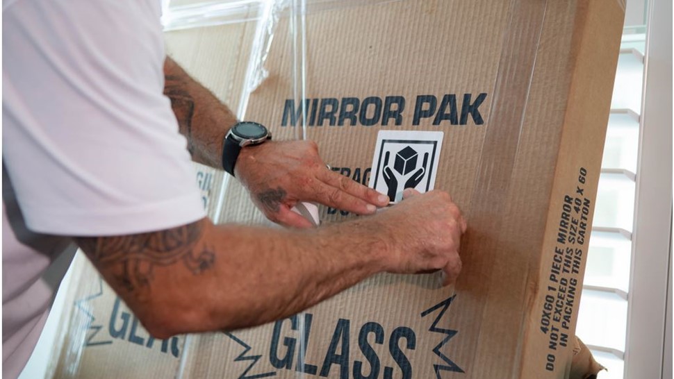 Mover placing a fragile sticker on a mirror pack box labeled glass for long-distance shipping.