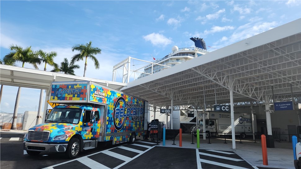 Colorful puzzle-pattern Solomon & Sons moving truck under a covered port terminal with palm trees and a cruise ship visible nearby.