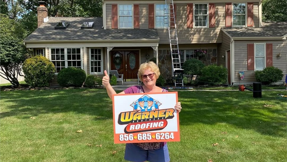 Smiling homeowner holding Warner Exteriors roofing sign in front yard