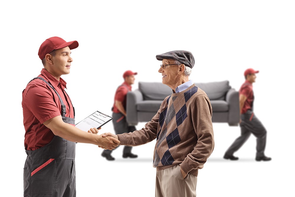 Elderly man shaking hands with a man with a clipboard as movers are carrying a couch in the background.
