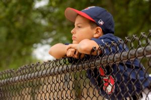 Boy wearing a Cleveland Indians uniform leans against a fence looking bored.