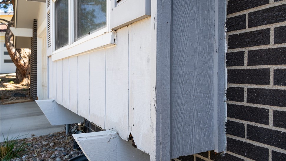 Close-up of chipped, worn Masonite siding and corner trim where it meets brick on a Longmont home