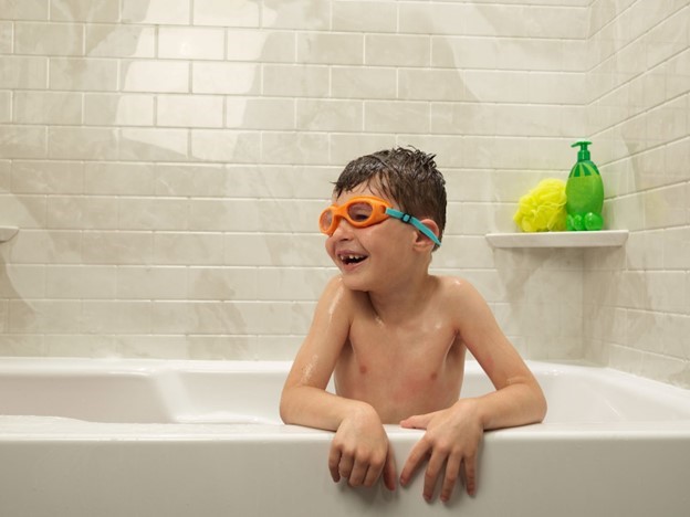 Photo of a smiling boy taking a bath with his arms over the side of a traditional tub and swimming goggles on.